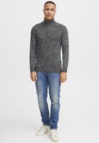 Dark gray textured pullover with a high collar and half zipper, paired with light blue jeans and white sneakers. Small logo on the hem.