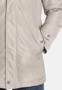 Chaqueta aislante beige con una textura suave, cierres a presión, bolsillos laterales y un diseño ligeramente ajustado en la cintura.