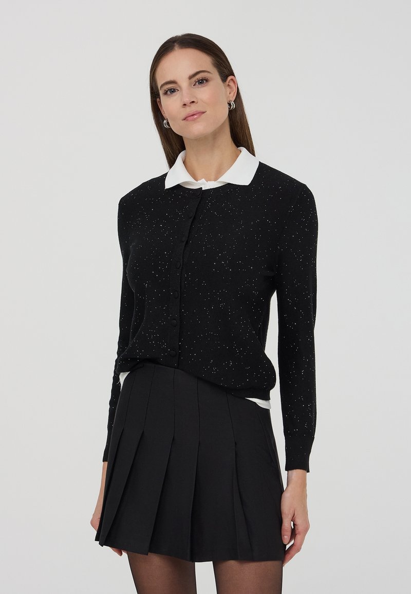 Donna che indossa un cardigan nero a pois con bottoni, una camicia bianca con colletto e una gonna plissettata nera, in piedi davanti a uno sfondo neutro.