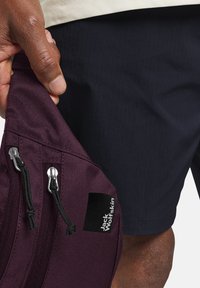 Zaino marrone con due cerniere argento, dettagli neri e etichetta "Jack Wolfskin", tenuto a mano contro pantaloni corti texture blu navy.
