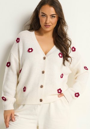 Vrouw draagt een witte gebreide cardigan met rode bloemapplicaties en knopen, gecombineerd met een witte broek, poseert tegen een effen achtergrond.