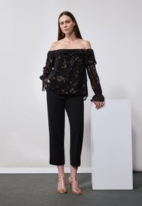 Top nero off-shoulder con maniche a volant e motivo floreale dorato, abbinato a pantaloni neri cropped e sandali dorati con cinturino. Piedistallo bianco sullo sfondo.