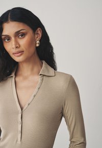 Camiseta de manga larga beige con textura acanalada y cuello con solapas, con detalles de botones y un sutil brillo. Pendientes redondos dorados distintivos.