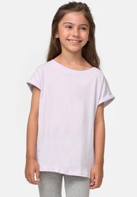 Ragazza sorridente con lunghi capelli castani, indossa una maglietta corta di colore viola chiaro e leggings grigi, in piedi su uno sfondo chiaro e semplice.