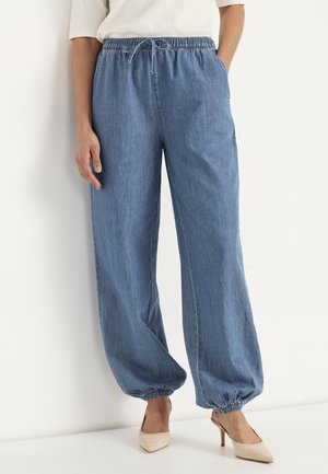Kvinde iført løstsiddende blå jeansbukser med elastisk talje og manchetter, kombineret med beige højhælede sko og en hvid top.