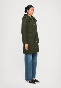 Femeie îmbrăcată într-un palton căptușit verde măsliniu cu glugă, blugi drepți de culoare albastră și pantofi kitten heels negri cu vârful ascuțit, stând în fața unui fundal alb.