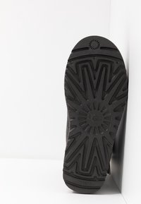 Semelle de botte noire avec un motif de semelle en zigzag profond et un dessin circulaire près du centre, placée debout contre un mur d’angle blanc.