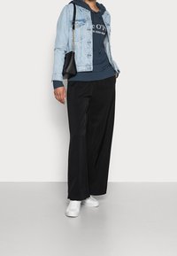 Chaqueta de mezclilla en azul claro sobre una sudadera con capucha navy con lettering, combinada con pantalones anchos negros y zapatillas blancas. Los acentos incluyen un pequeño bolso negro.