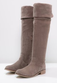 Botas de ante taupe hasta la rodilla con tacón plano, cierre de cremallera lateral y diseño de puño doblado. Textura suave con detalles de herrajes mínimos.