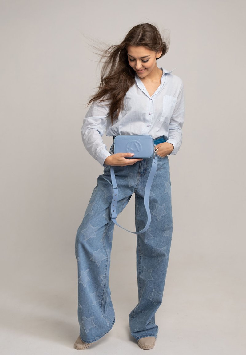 Borsa a tracolla blu in ecopelle con forma rettangolare e motivo a stelle, indossata su una camicia oversize azzurro chiaro e jeans a gamba larga.