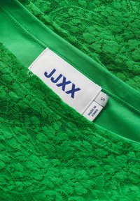 Tissu en dentelle verte avec une doublure verte lisse. Présente une étiquette blanche avec un texte bleu — nom de la marque « JJXX » et taille « S ». 