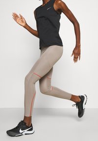 Henkilö, joka on pukeutunut mustaan hihattomaan paitoihin ja beigeihin legginsseihin, joissa on punaista ommelta, juoksee mustat ja valkoiset Nike-juoksukengät jalassa.