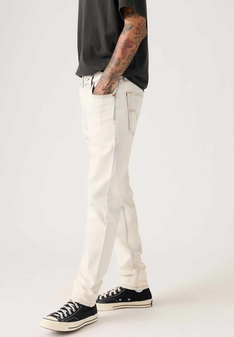 Jean en denim clair à coupe slim, avec des coutures contrastantes et un subtil rétrécissement vers les chevilles, associé à des baskets noires.