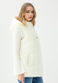Cremfarbener Puffer-Weste mit Kapuze, ausgestattet mit Druckknöpfen, zwei Fronttaschen und einer glatten Textur, getragen über einem hellen Langarmoberteil.