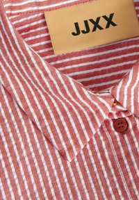 Rood en wit gestreefd overhemd met kraag, voorzien van een klein bruin etiket met de tekst "JJXX." Heeft twee bruine knoppen op de knoopsluiting en een gestructureerde stof.