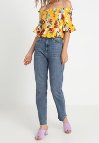 Top amarillo con hombros descubiertos, estampado floral y borde con volantes, combinado con vaqueros azules de talle alto y mules de tacón bloque lavanda.