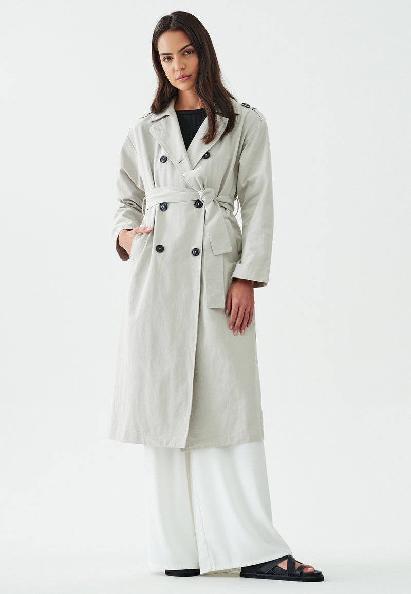 Helehall trench coat kahekordsete nuppudega, vöödiga talje ja küljetaskudega. Mudel kannab laiad valged püksid ja musti sandleid.