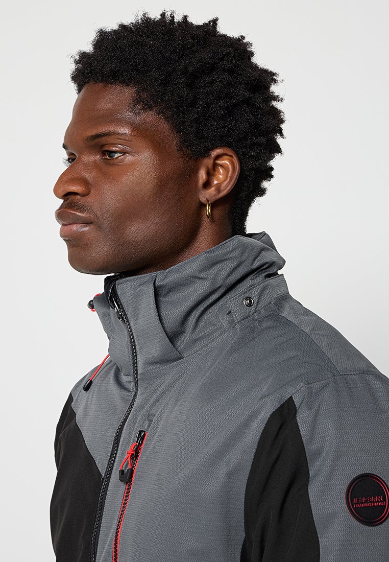 Hombre con pelo rizado corto que lleva una chaqueta gris y negra con detalles en rojo en el zipper y un aro dorado en la oreja, mirando hacia la izquierda sobre un fondo liso.