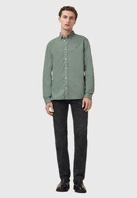 Groene button-up shirt met lange mouwen, kraag en slim fit, gecombineerd met donkere jeans en zwarte schoenen, staand tegen een neutrale achtergrond.