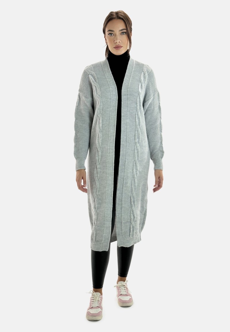 Grå stickad cardigan med flätstickade mönster, långa ärmar och öppen framdesign, kombinerad med svarta leggings och pastellfärgade sneakers.