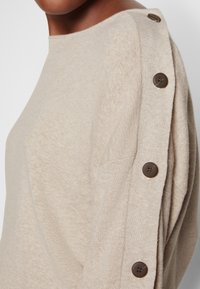 Pull en tricot beige avec un col bateau et des boutons marron foncé le long de l'épaule et du haut de la manche.