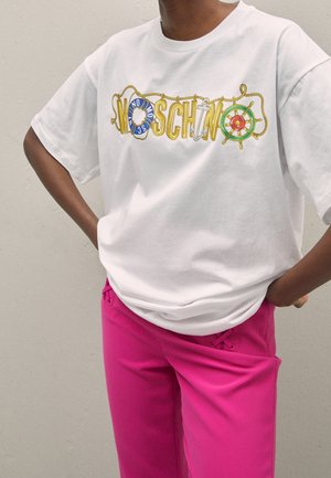 Osoba ubrana w biały t-shirt z morską grafiką i logo "MOSCHINO" oraz jasnoróżowe spodnie, z rękami opartymi na biodrach.