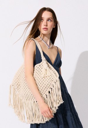 Junge Frau in einem dunkelblauen ärmellosen Kleid trägt eine große cremefarbene Makramee-Umhängetasche mit Fransen, mit leicht im Wind wehenden langen Haaren.