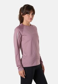 Woman with brown hair in a ponytail wearing a mauve long-sleeve athletic top and black leggings, standing against a plain light background.