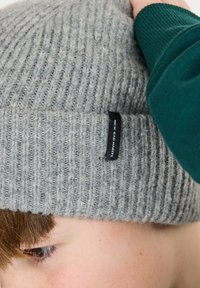Grijze geribbelde gebreide beanie met een omgeslagen rand en een klein zwart label met de tekst "NEW BAD HABITS." Zachte textuur met een warme uitstraling.