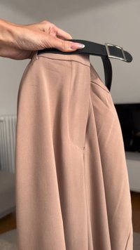 Bež krilo iz tkanine, ki ga drži črn pas s srebrno zaponko. Krilo ima mehak teksturo in subtilno nabiranje na pasu.