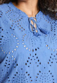 Blusa blu ricamata con motivi a intaglio, scollatura rotonda e chiusura a fiocco con nappine. Realizzata in tessuto leggero e testurizzato.