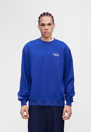 Junger Mann mit geflochtenem Haar, der einen blauen Red Bull Racing Sweatshirt und dunkle Hosen trägt, steht vor einem schlichten weißen Hintergrund.