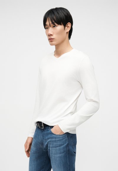 Nuori mies, joka pukeutunut valkoiseen pitkähihainen paitaan ja sinisiin farkkuihin, toinen käsi taskussa, seisomassa yksiväristä taustaa vasten.