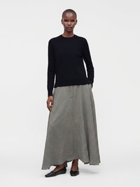 Femme portant un haut à manches longues noir, une jupe grise longueur cheville et des chaussures plates noires, debout devant un fond clair uni.