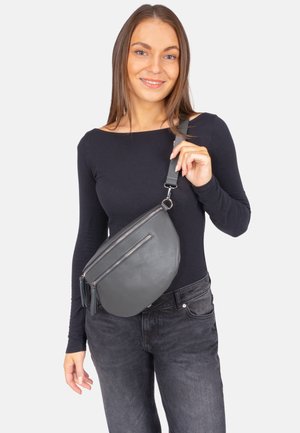 Junge Frau in schwarzem Langarmshirt und Jeans hält eine graue Leder-Umhängetasche mit Reißverschlusstaschen vor weißem Hintergrund.