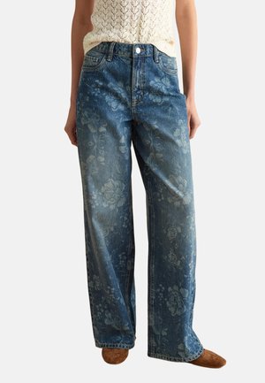 Jeans de mezclilla azul de pierna ancha con un sutil estampado floral, combinados con un top sin mangas de punto color crema y zapatos marrones.