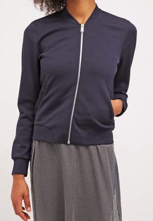 Woman wearing a dark navy zip-up bomber jacket with ribbed cuffs and collar, paired with a gray pleated skirt.