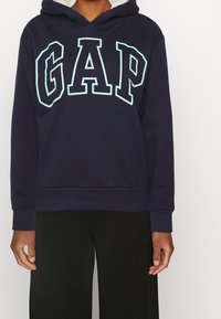 Sweat à capuche en coton bleu marine avec un grand logo "GAP" bleu clair sur le devant, doté d'une capuche à cordon de serrage et de poignets et ourlet côtelés.