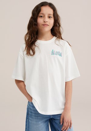 Dziewczyna z długimi kręconymi brązowymi włosami, ubraną w białą, oversize koszulkę z napisem „ALOHA” i niebieskie dżinsy, stojąca z jedną ręką w kieszeni.