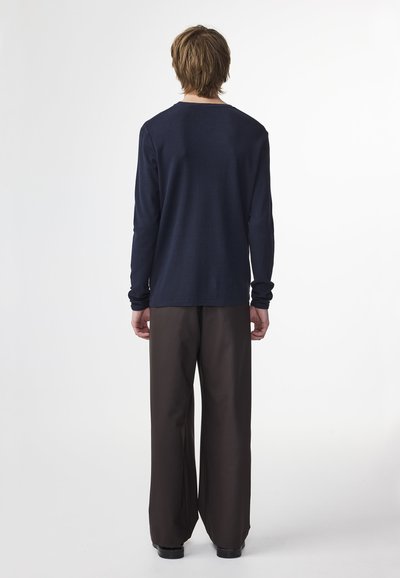 Mann steht mit dem Rücken zur Kamera, trägt ein marineblaues Langarmhemd, dunkle weitgeschnittene Hose und schwarze Schuhe vor einem schlichten weißen Hintergrund.
