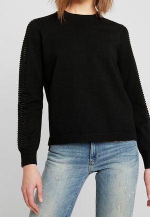 Femme portant un pull noir à manches longues avec des détails côtelés sur les manches et un jean bleu clair déchiré.