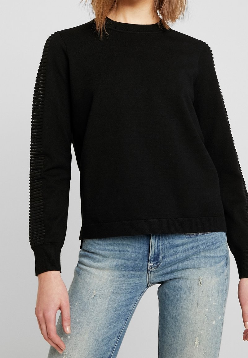 Femme portant un pull noir à manches longues avec des détails côtelés sur les manches et un jean bleu clair déchiré.