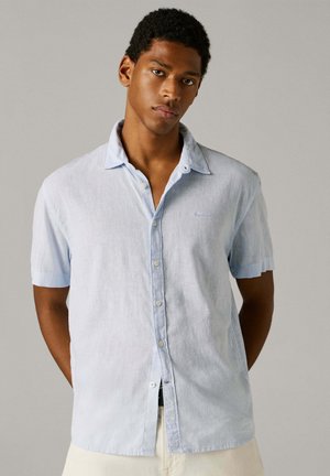 Νέος άνδρας με κοντά και σγουρά μαλλιά, που φοράει ένα ανοιχτό γαλάζιο πουκάμισο με κοντά μανίκια και κουμπιά, μπροστά σε ένα απλό μπεζ φόντο.