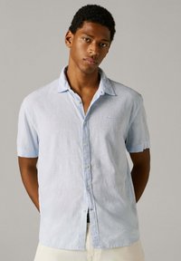 Νέος άνδρας με κοντά και σγουρά μαλλιά, που φοράει ένα ανοιχτό γαλάζιο πουκάμισο με κοντά μανίκια και κουμπιά, μπροστά σε ένα απλό μπεζ φόντο.