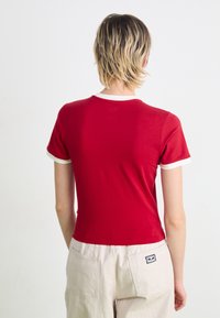 Raudona trumpa rankovė t-shirt su balta apykaklės ir rankovių apdaila, pagaminta iš minkštos medžiagos. Nugaroje matomas prigludęs dizainas.