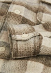 Țesătură de cămașă din flanel cu carouri bej și maron, având un manșon cu nasturi, din lână moale și texturată, cu un model de carouri proeminent.