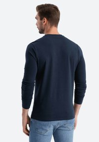 Tummansininen pitkähihaisen paita pehmeästä kankaasta, jossa on kapeat hihat ja pyöreä kaula-aukko, yhdistettynä vaaleansinihin farkkuihin, nähtynä takaa.