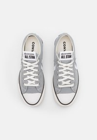 Une paire de baskets Converse All Star basses grises avec des embouts en caoutchouc blanc et des lacets blancs sur un fond blanc.