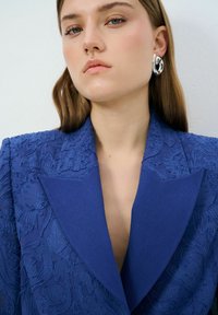 Blazer bleu avec un broderie florale texturée, des revers aigus et un décolleté plongeant. Le mannequin porte des boucles d'oreilles circulaires en argent. Finition en tissu lisse.