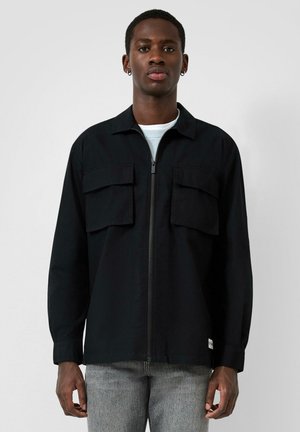 Jeune homme à la peau foncée portant des boucles d'oreilles argentées en forme d'anneau, une veste noire zippée sur un T-shirt blanc et un jean gris, debout devant un fond uni.